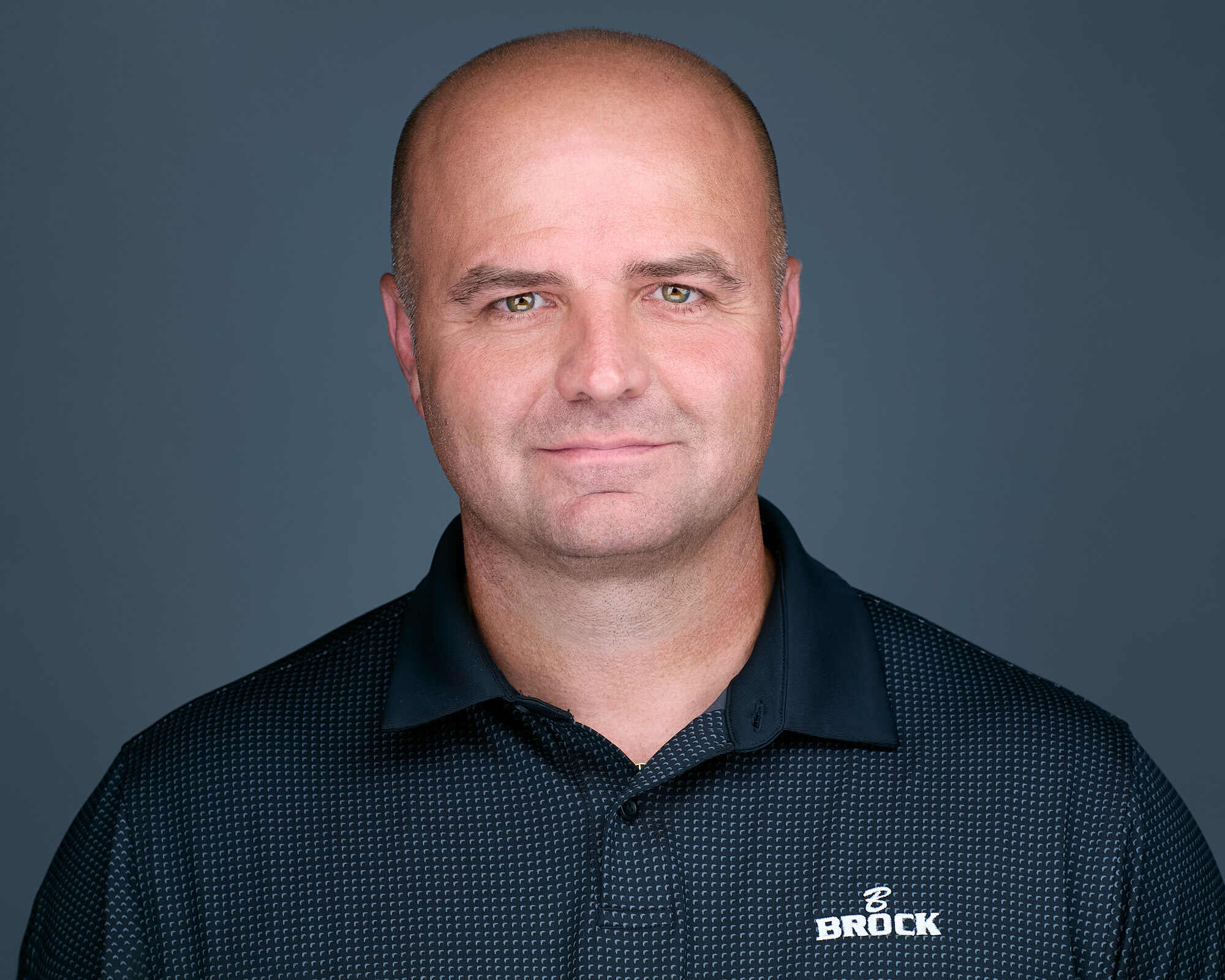 Headshot of a man with a shaved head, wearing a black collared shirt with a subtle checkered pattern and the 'Brock Builders' logo on the left side. He has a neutral expression, light skin, and is set against a plain grey background.