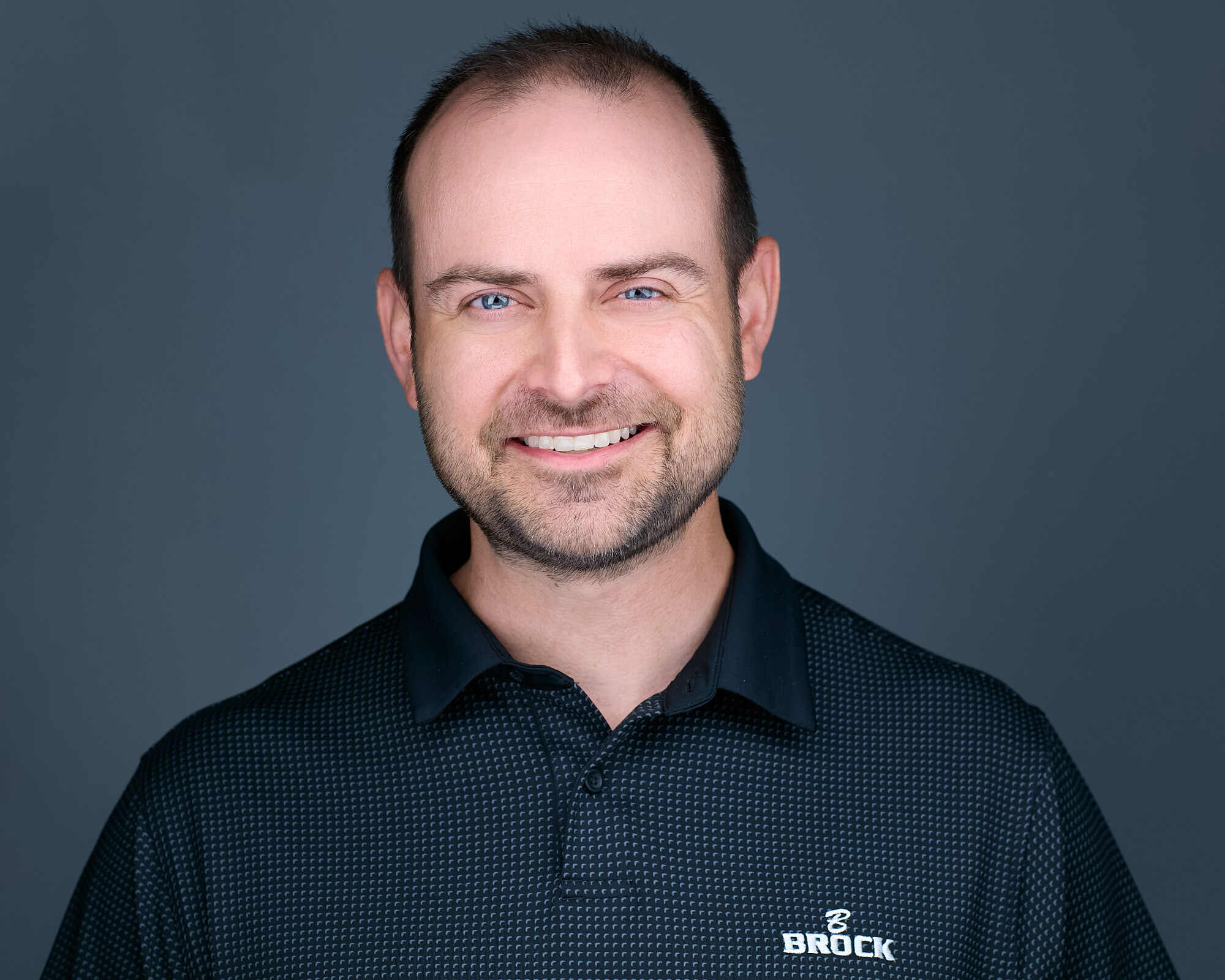 Headshot of a man with short dark hair and a trimmed beard, wearing a black collared shirt with a subtle checkered pattern and the 'Brock Builders' logo on the left side. He has a friendly expression, light skin, and is set against a plain grey background.