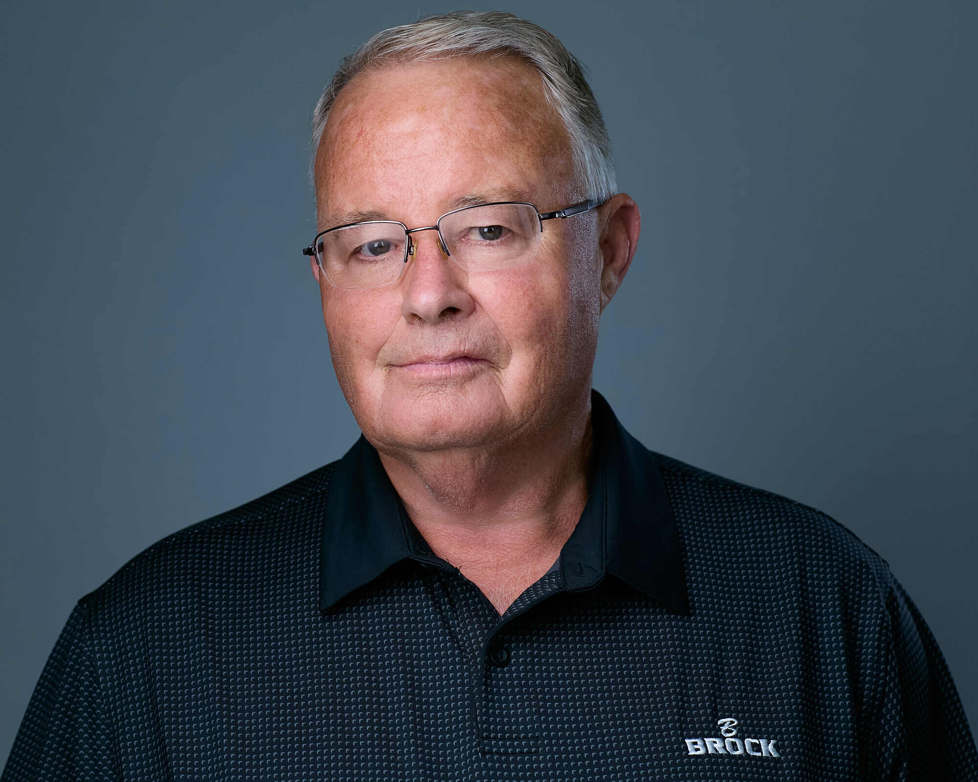 Headshot of an older man with short grey hair, wearing glasses and a black collared shirt with a subtle checkered pattern and the 'Brock Builders' logo on the left side. He has a neutral expression, light skin, and is set against a plain grey background.