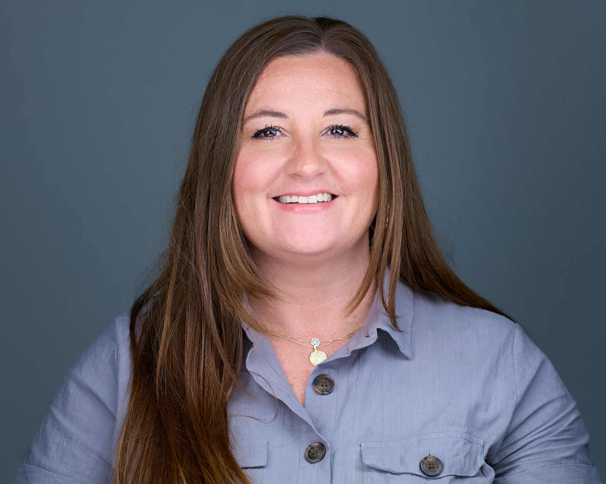 Headshot of a woman with long straight brown hair, wearing a light grey collared shirt with large buttons and a gold necklace with a pendant. She has a friendly smile, light skin, and is set against a plain grey background.