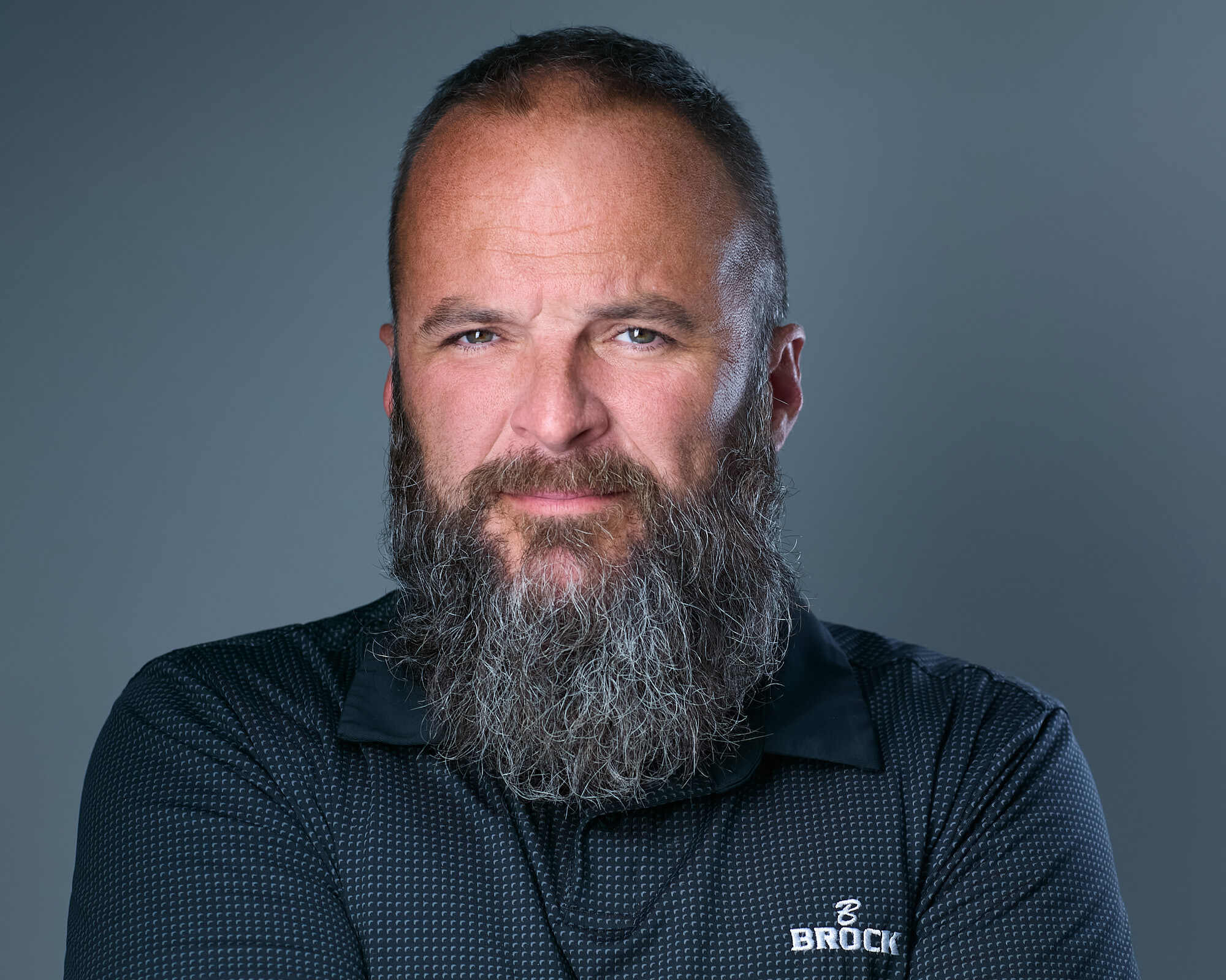 Headshot of a middle-aged man with a long, thick grey beard and short hair, wearing a black collared shirt with a subtle checkered pattern and the 'Brock Builders' logo on the left side. He has a confident expression, light skin, and is set against a plain grey background.