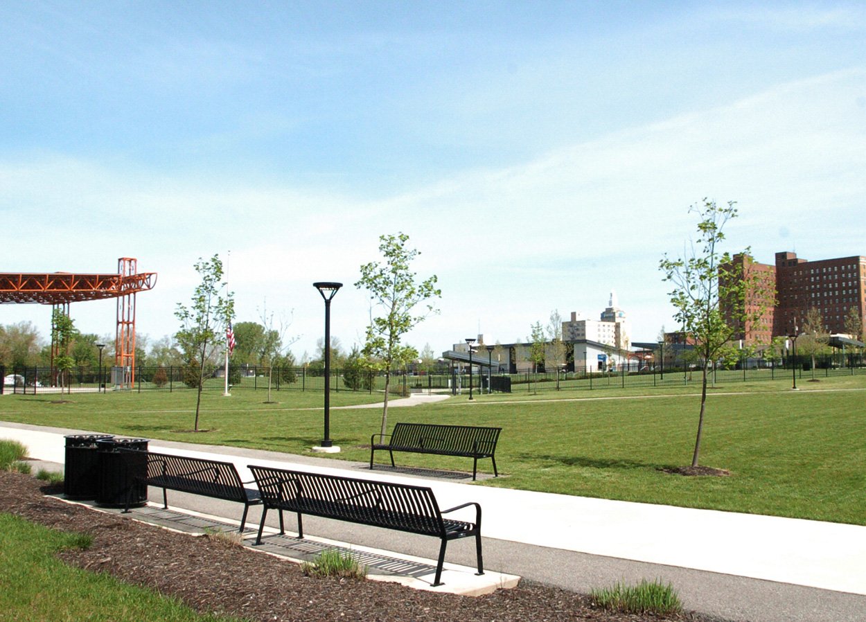 A park with green grass, young trees, and paved walkways. The park features several black metal benches and lampposts. In the background, there is an orange metal structure, possibly a stage or pavilion, and buildings from a nearby city. The sky is clear and blue.