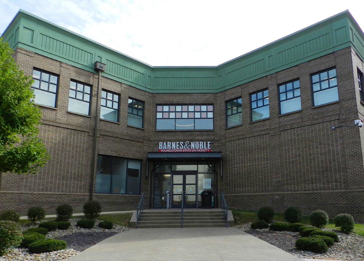 Exterior of the Barnes & Noble Youngstown State University building. The building is made of brown brick with large windows and a green cornice. A sign above the entrance reads 'Barnes & Noble Youngstown State University.' There are steps leading up to the entrance, and the area is landscaped with small bushes and stones.