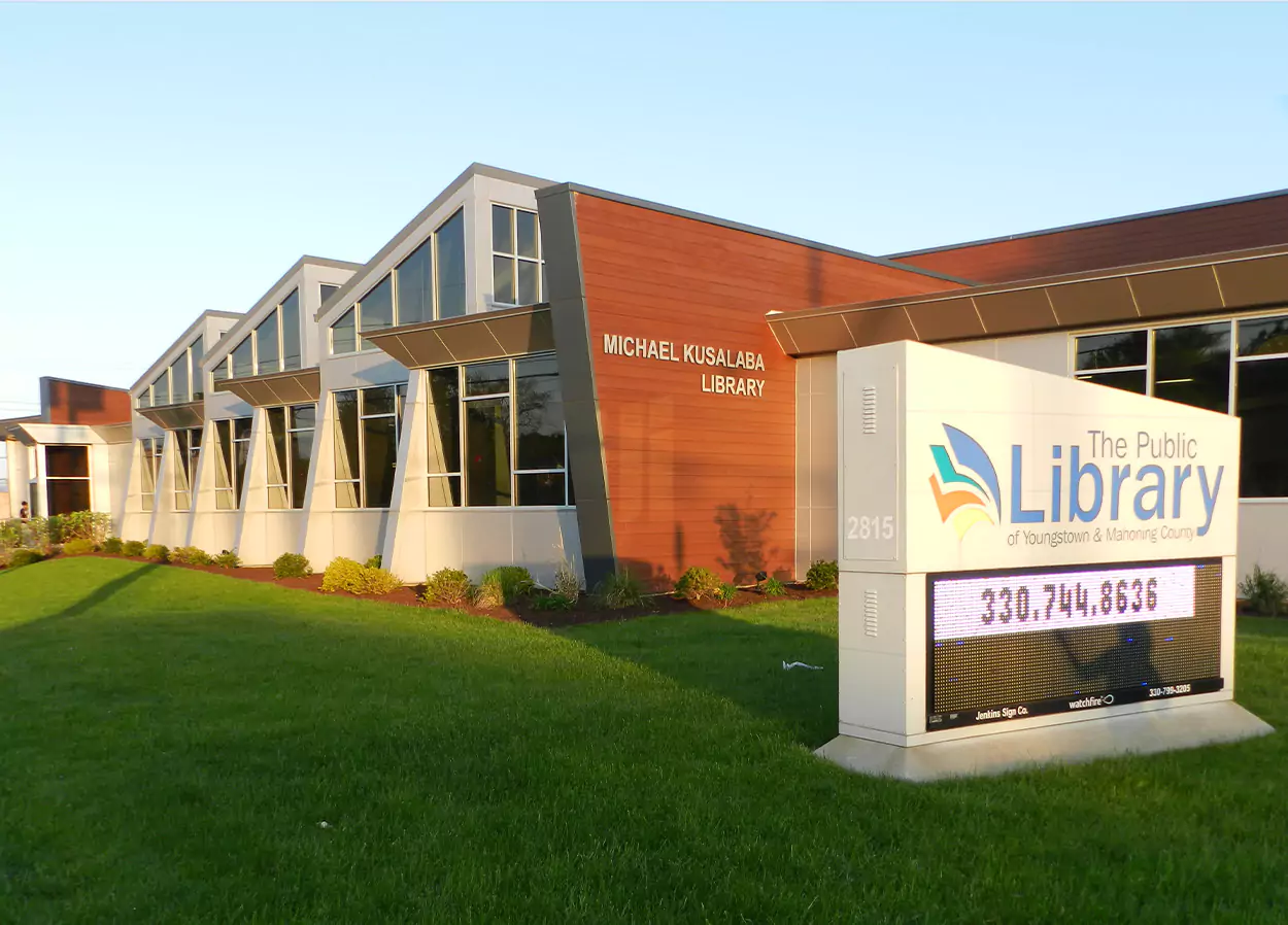 Exterior of the Michael Kusalaba Library, a modern building with large windows and a mix of wood and metal paneling. A sign in front of the building reads 'The Public Library of Youngstown & Mahoning County' with a phone number displayed. The surrounding area is landscaped with grass and small bushes, and the sky is clear and blue.