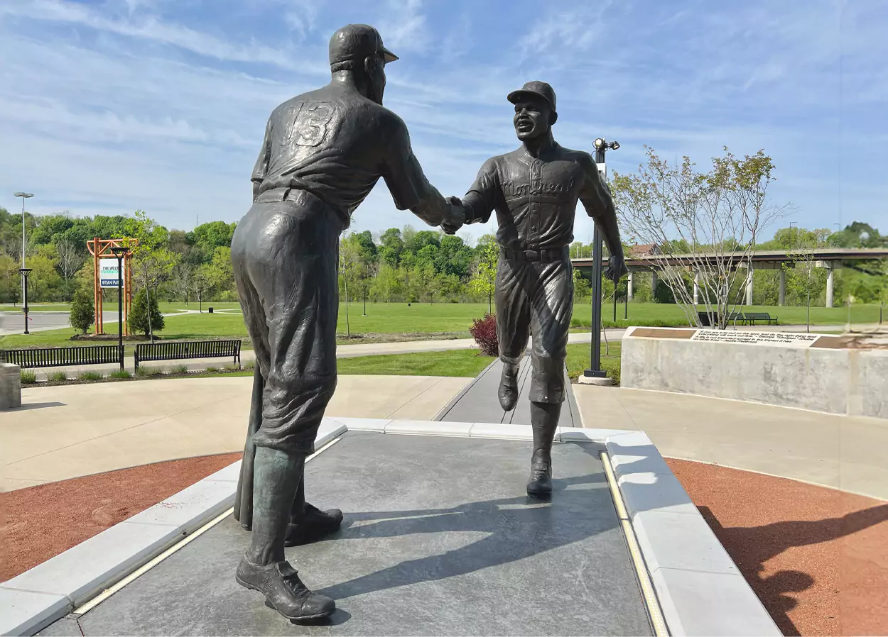 A statue of two baseball players shaking hands on a concrete platform. The player on the left has the number 13 on his back, and both players are dressed in vintage baseball uniforms. The background includes a park with green trees, benches, and a sign that reads 'Wean Park'.