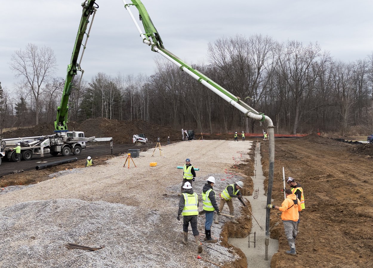 Construction site with workers in safety vests and helmets working on a gravel foundation. A large green concrete pump truck is in operation, pouring concrete. The background features bare trees and an overcast sky. Equipment and construction materials are scattered around the site.